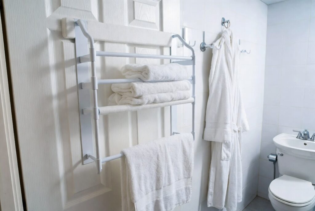 Over-the-door towel rack with padded brackets on bathroom door holding white towels and robe. No-drill, no-damage solution for apartments and small bathrooms.