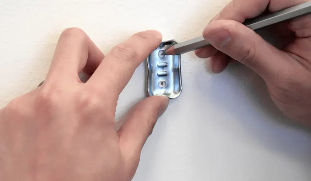Hands holding a metal mounting bracket against a white wall while marking drill hole locations with a pencil