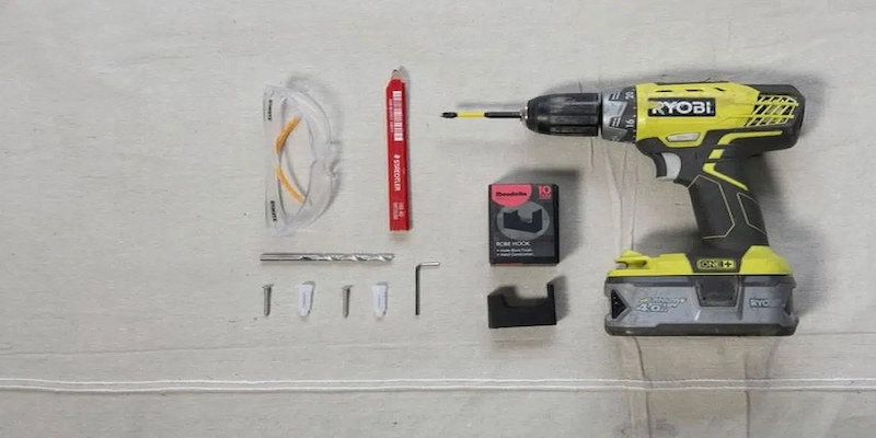 Flat lay of tools for installing a robe hook: Ryobi cordless drill, safety glasses, red carpenter pencil, drill bit, screws, wall anchors, Allen key, and robe hook mounting hardware arranged on a light fabric surface