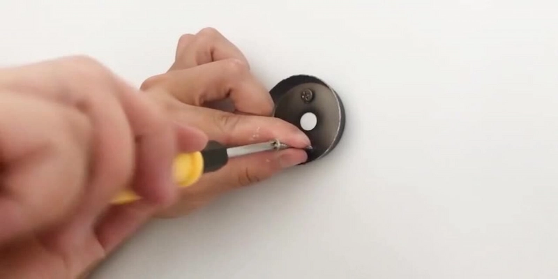 Hands using a yellow-handled screwdriver to tighten a screw on a round black mounting plate for a robe hook on a white wall