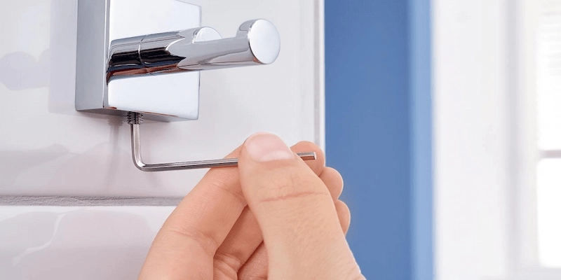 Close-up of a hand using an Allen wrench to tighten a screw on a chrome bathroom towel hook mounted on white tile wall